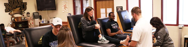 In the athletic training facility Professor Samantha Wilson sits between two students on the cushioned station while a student wraps her ankle.