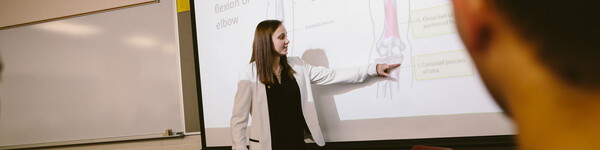 Professor Tamra Llewellyn in the front of a class pointing to points at an illustration of bones in an arm.